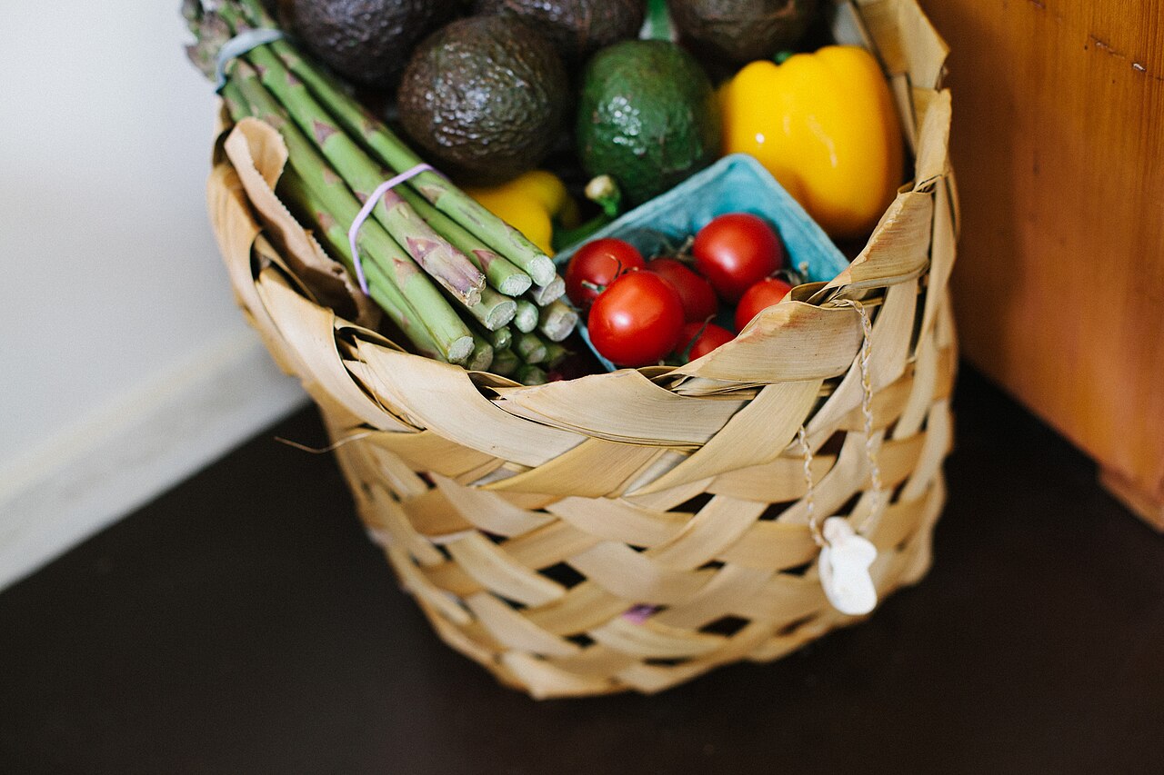 A woven basket of asparagus, avocados, tomatoes and a yellow pepper